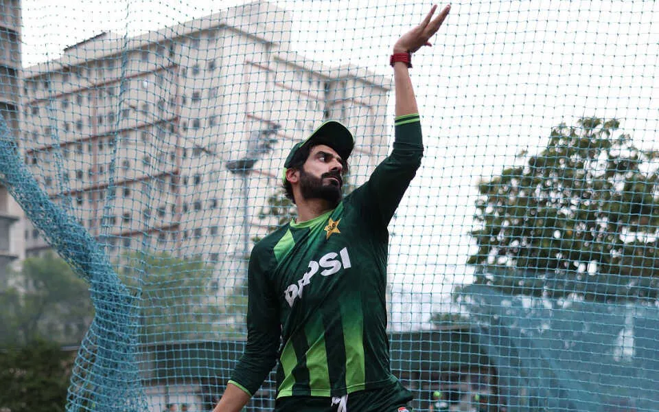 A focused cricketer holding a bat, ready to play on an artificial turf field. The backdrop includes a colorful advertisement and a netted area.